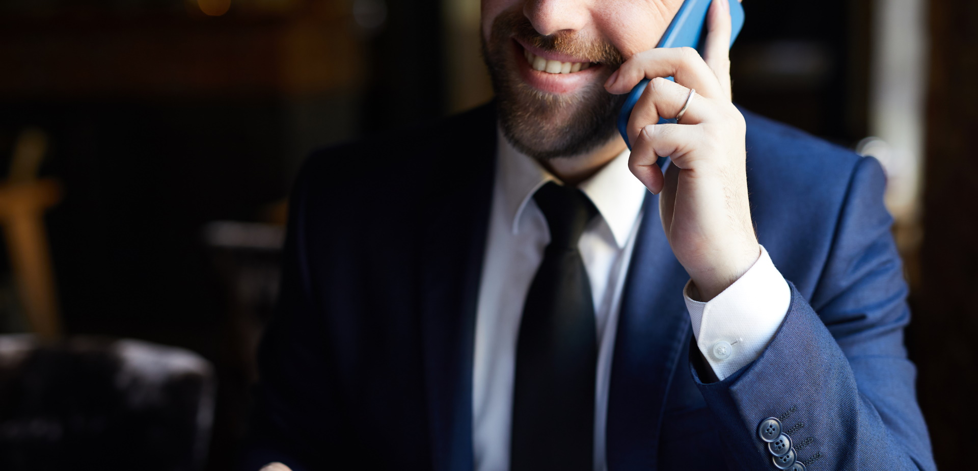 Close-up of positive bearded businessman in elegant suit with coffee cup calling on mobile phone and smiling while sitting at table